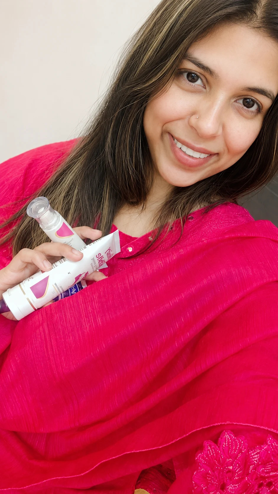 Zarqoon Saeed holding a white skincare tube against a vivid hot-pink dupatta, warm smile, hair down